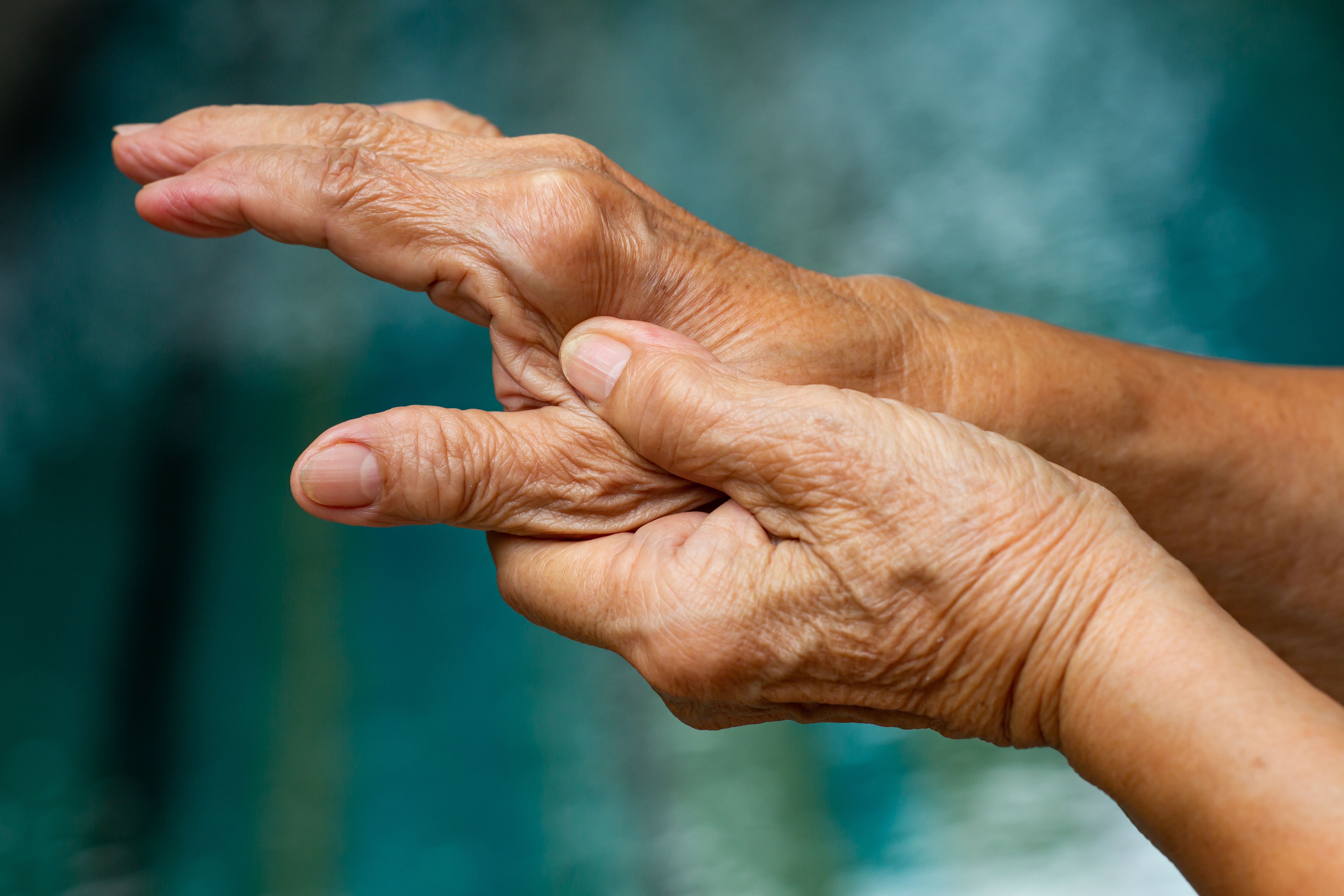 Close-up of an older person's hands, with the thumb of one hand pressing into the palm of the other, against a blurred teal background.  | Image Credit: © eyepark - stock.adobe.com