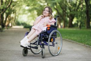 Portrait of pleasant young woman with spinal muscular atrophy smiling on camera among green summer park. Female person who using wheelchair. Concept of people with disability. | Image Credit: © sofiko14 - stock.adobe.com