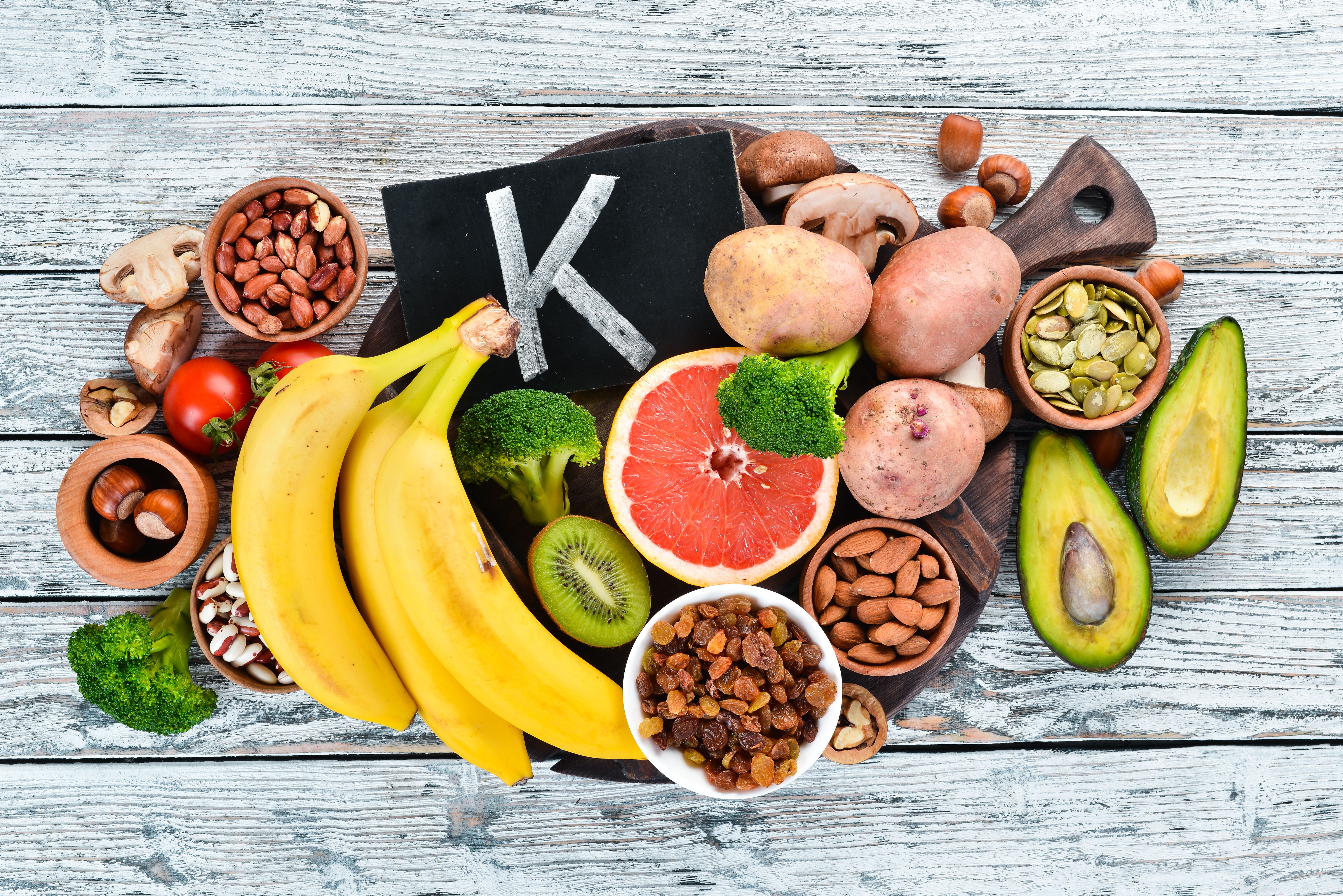 Foods containing natural potassium. K: Potatoes, mushrooms, banana, tomatoes, nuts, beans, broccoli, avocados. Top view. On a white wooden background. | Image Credit: © Yaruniv-Studio