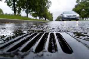 Close-up of storm drain during rainfall, car driving in background on wet road. © Moopingz - stock.adobe.com