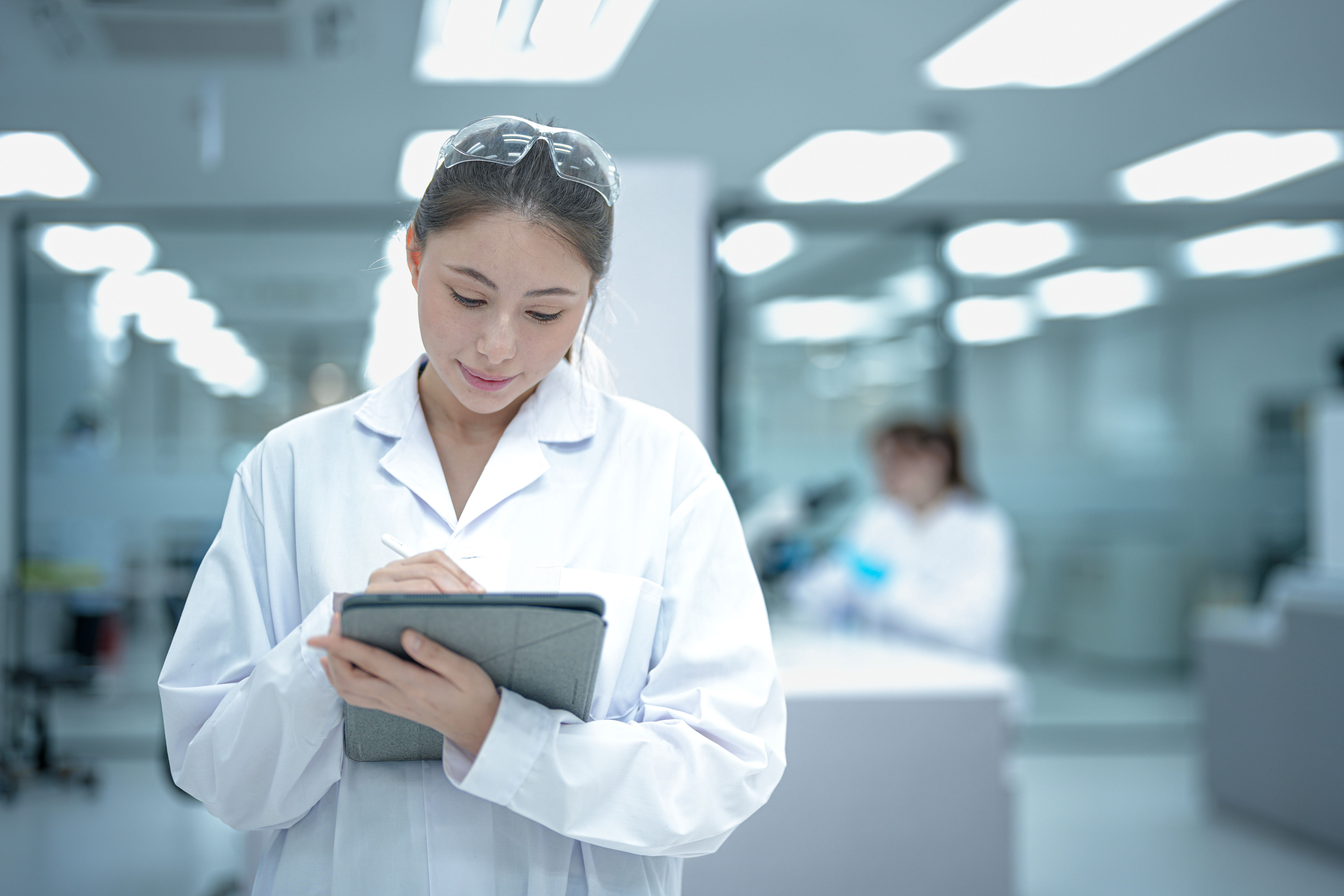 A confident young female scientist in a lab coat and goggles stands with arms crossed in a modern laboratory, representing the bright future of women in STEM careers and research. | Image Credit: © ultramansk - stock.adobe.com.