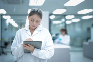 A confident young female scientist in a lab coat and goggles stands with arms crossed in a modern laboratory, representing the bright future of women in STEM careers and research. | Image Credit: © ultramansk - stock.adobe.com.