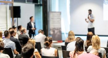 Business and entrepreneurship symposium. Speaker giving a talk at business meeting. Audience in conference hall. Rear view of unrecognized participant in audience. | Image Credit: © kasto - stock.adobe.com 