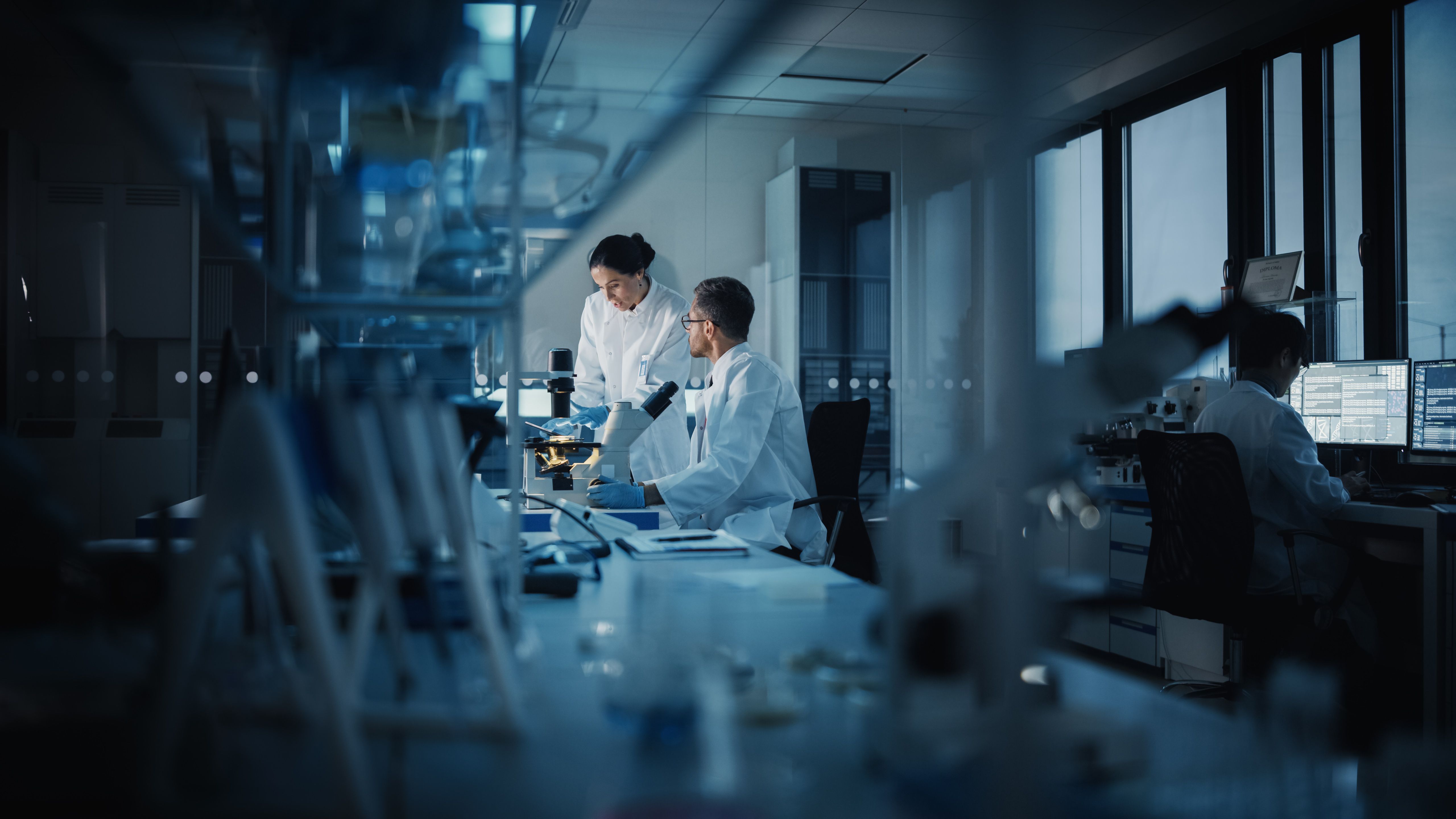 Modern Medical Research Laboratory: Two Scientists Working, Using Digital Tablet, Analyzing Test, Talking. Advanced Scientific Pharmaceutical Lab for Medicine, Biotechnology Development. Evening Time | Image Credit: © Gorodenkoff - stock.adobe.com