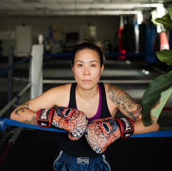 Stacy Leung stands in a boxing ring with her two arms up on the ropes with boxing globes on.