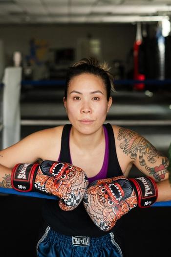 Stacy Leung stands in a boxing ring with her two arms up on the ropes with boxing globes on.