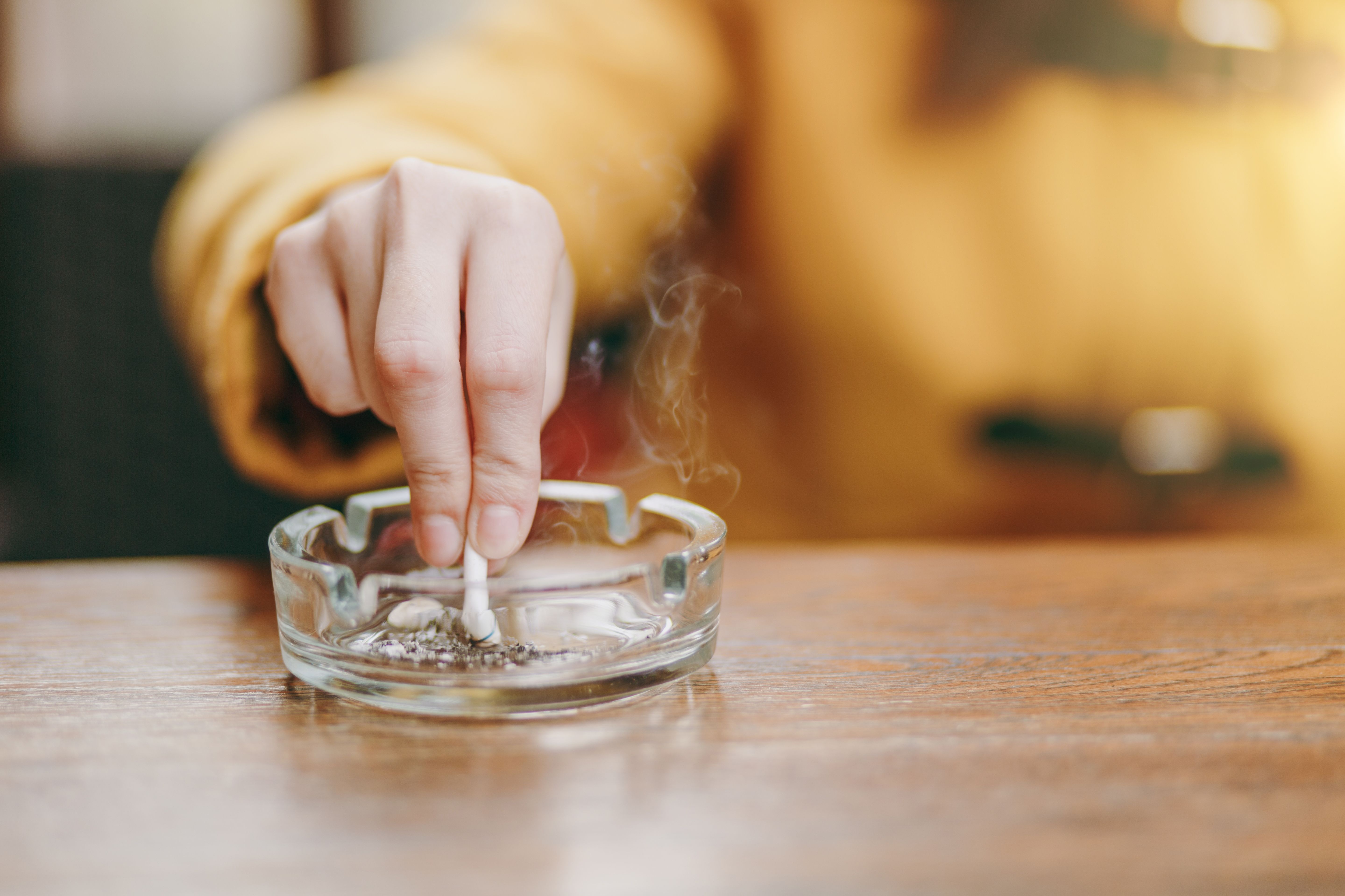 Focus on caucasian young woman hand putting out cigarette on glass ashtray on wooden table, cigarette butt, smoking is dying. Quit smoking. Health concept. Close up photo. | Image credit: ViDi Studio - © - stock.adobe.com
