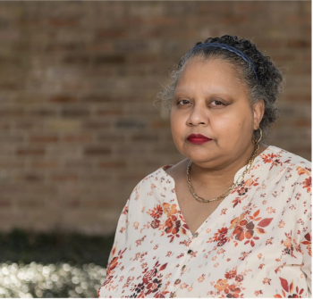 Image of a woman with curly gray hair with white and peach-colored floral blouse, looking pensive at the camera. 
