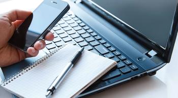 Image of a person holding a smartphone besides their laptop and a notepad with a pen on top.