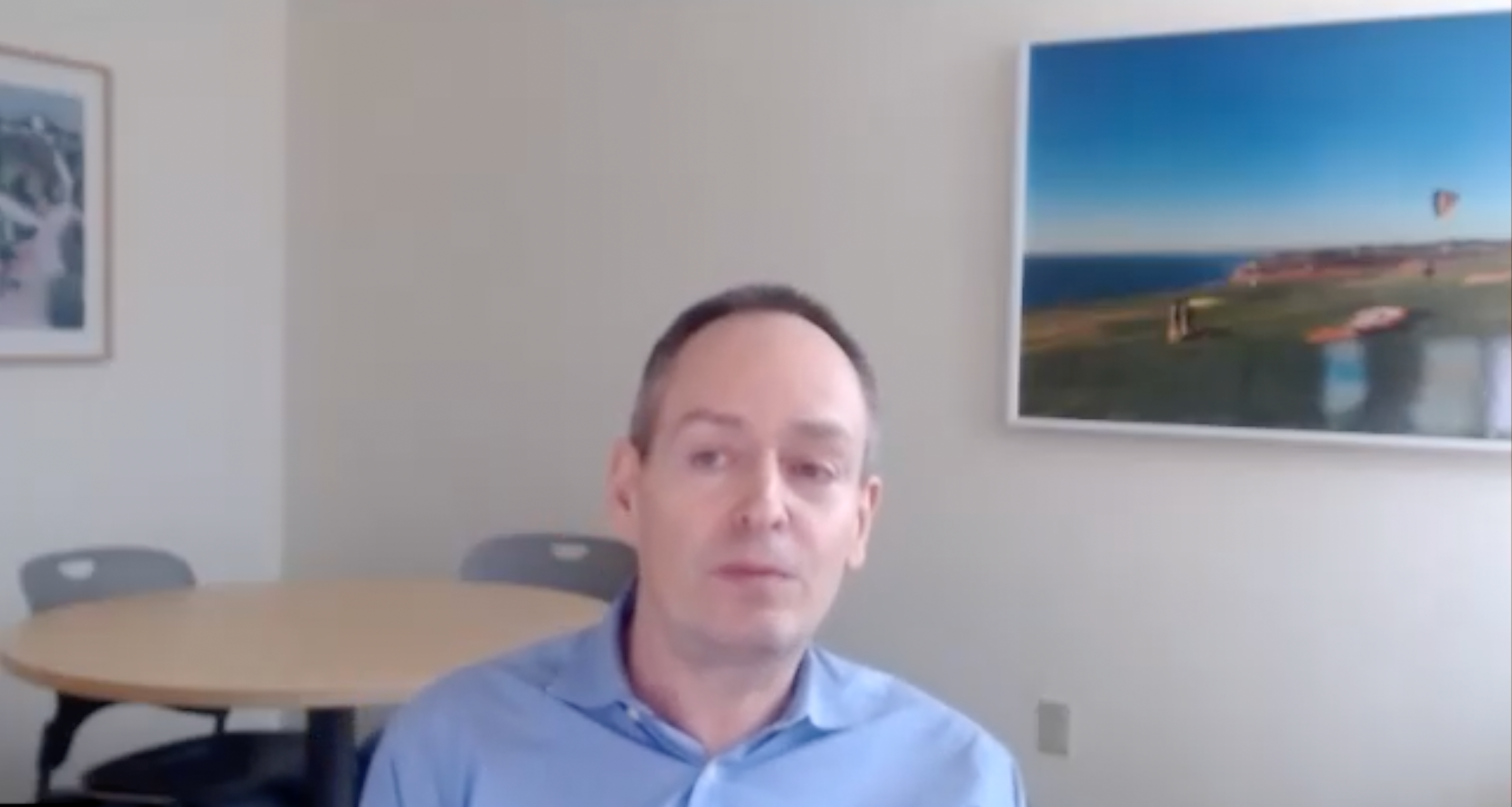 Dr. Patrick Ott of Dana-Farber Cancer Institute wearing a blue button up shirt, sitting in a room with two nature paintings.
