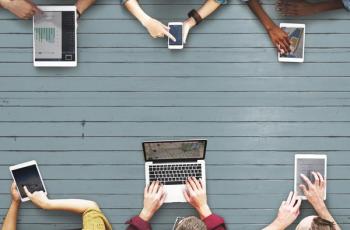 Image of six people at a table looking at six different technology devices, including phones, tablets and laptops.