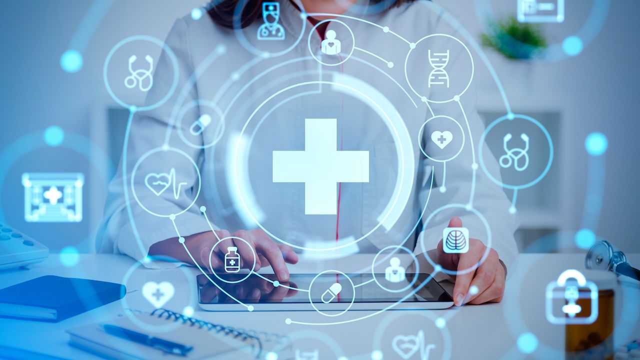 Female doctor sitting with table and various health care icons | Image credit: stock.adobe.com - ImageFlow