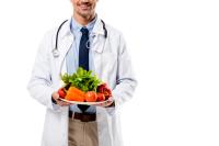 man wearing white doctor coat with stethoscope draped around neck holding a tray of fruit and vegetables | image credit: @Lighthouse Studios stock.adobe.com