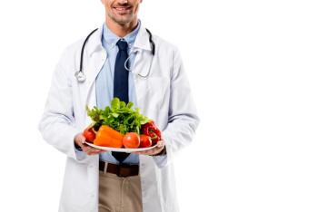 man wearing white doctor coat with stethoscope draped around neck holding a tray of fruit and vegetables | image credit: @Lighthouse Studios stock.adobe.com