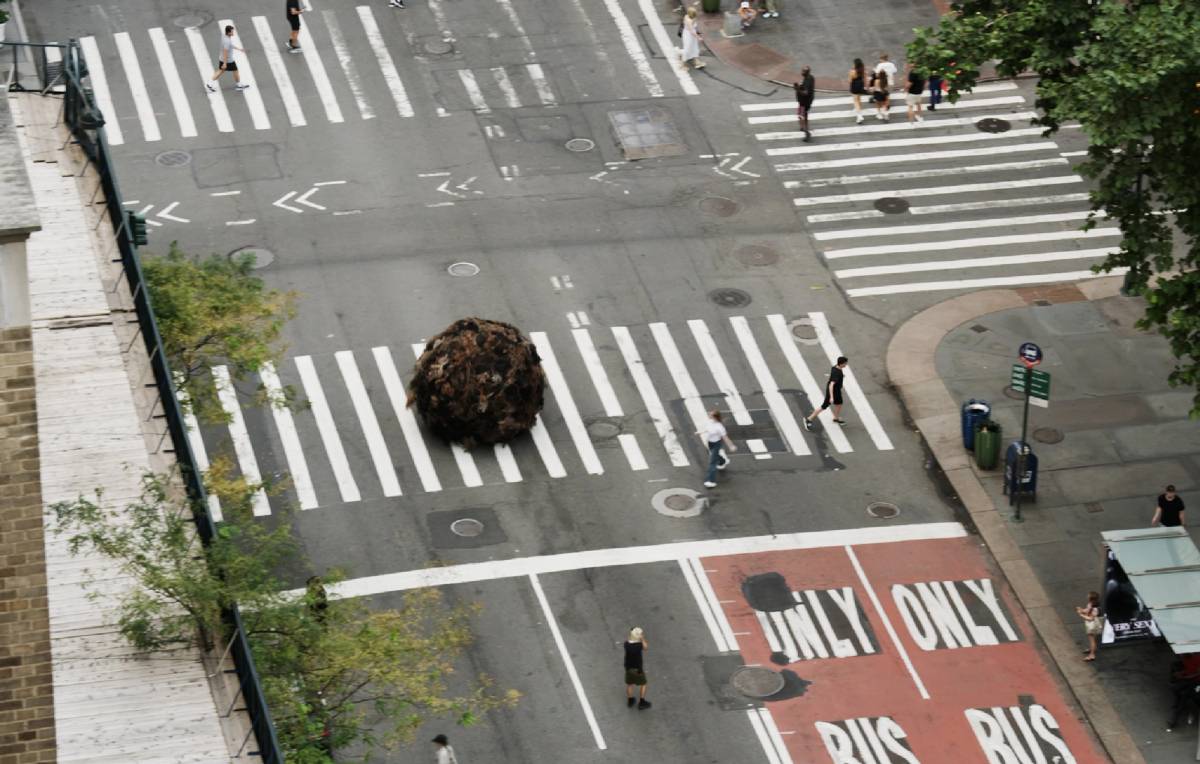 Aerial view of K18's giant "Tumblehair" installation sitting in a New York City crosswalk, with pedestrians crossing the street around it and a bus-only lane visible nearby.