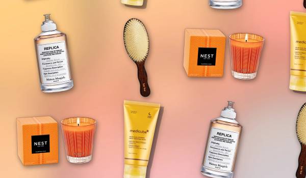 Flat-lay grid of beauty products on a warm gold-to-pink gradient background — a Maison Margiela Replica 'Coffee Break' perfume bottle, a Bur Bur wooden hairbrush with cream bristles, a Nest New York Pumpkin Chai candle with its striped orange box and a golden Medicube Kojic Acid Turmeric night mask tube