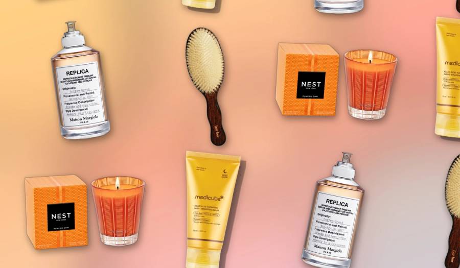 Flat-lay grid of beauty products on a warm gold-to-pink gradient background — a Maison Margiela Replica 'Coffee Break' perfume bottle, a Bur Bur wooden hairbrush with cream bristles, a Nest New York Pumpkin Chai candle with its striped orange box and a golden Medicube Kojic Acid Turmeric night mask tube
