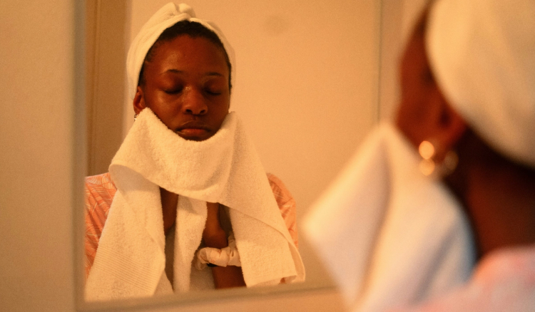 Woman with a towel wrapped around her hair pats her face dry with a white towel, reflected in a bathroom mirror.