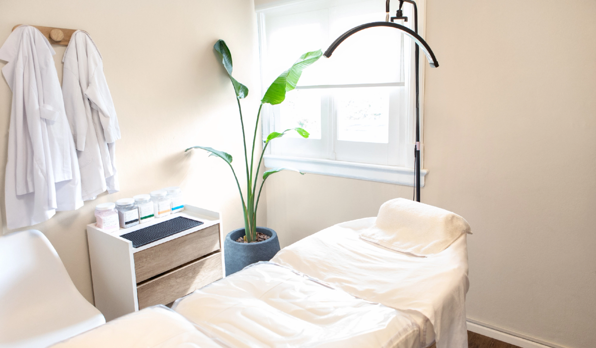 Bright treatment room with a white facial bed, overhead light, skin care supplies and a potted plant beside a window.