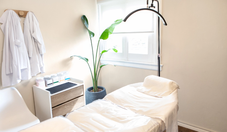 Bright treatment room with a white facial bed, overhead light, skin care supplies and a potted plant beside a window.
