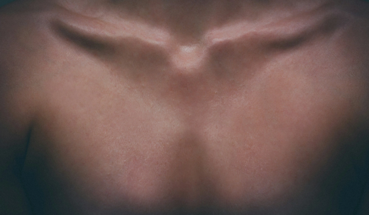 Close-up of a person’s bare upper chest and collarbones in soft, moody light.