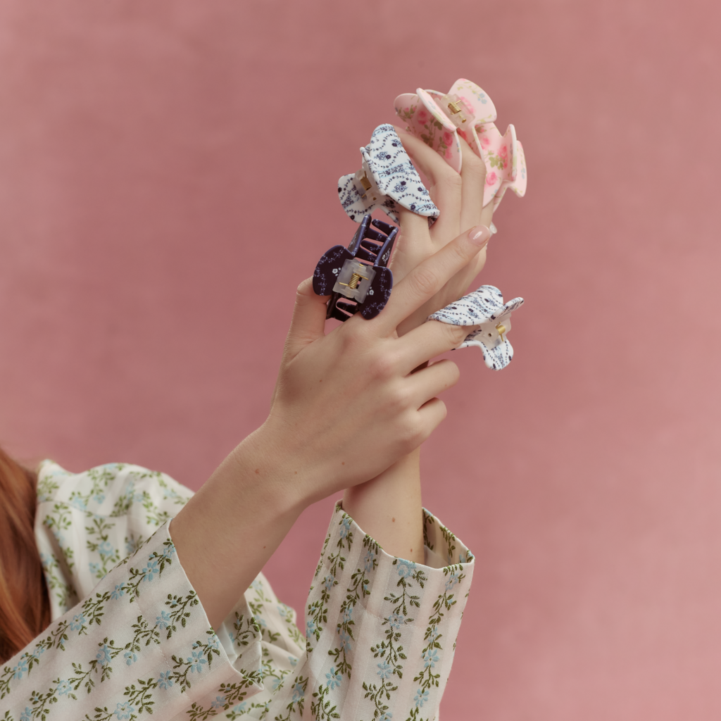 Model holding multiple patterned claw clips from the Wet Brush x Hill House Home collection in floral and gingham prints against a pink background.