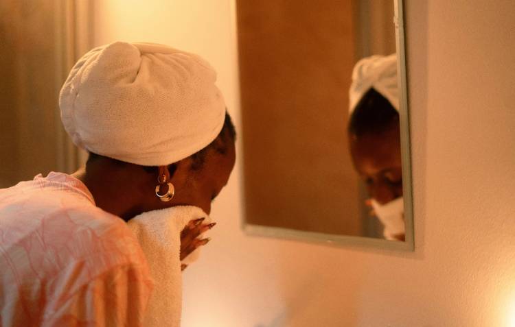 Woman with a towel wrapped around her hair drying her face in front of a mirror