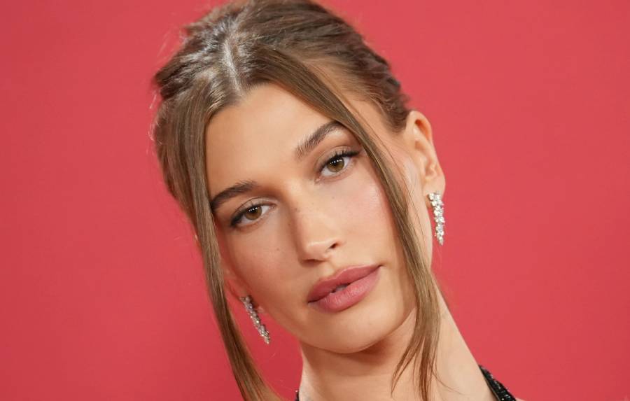Hailey Bieber wears soft glam makeup and a slicked-back updo with face-framing pieces against a red backdrop