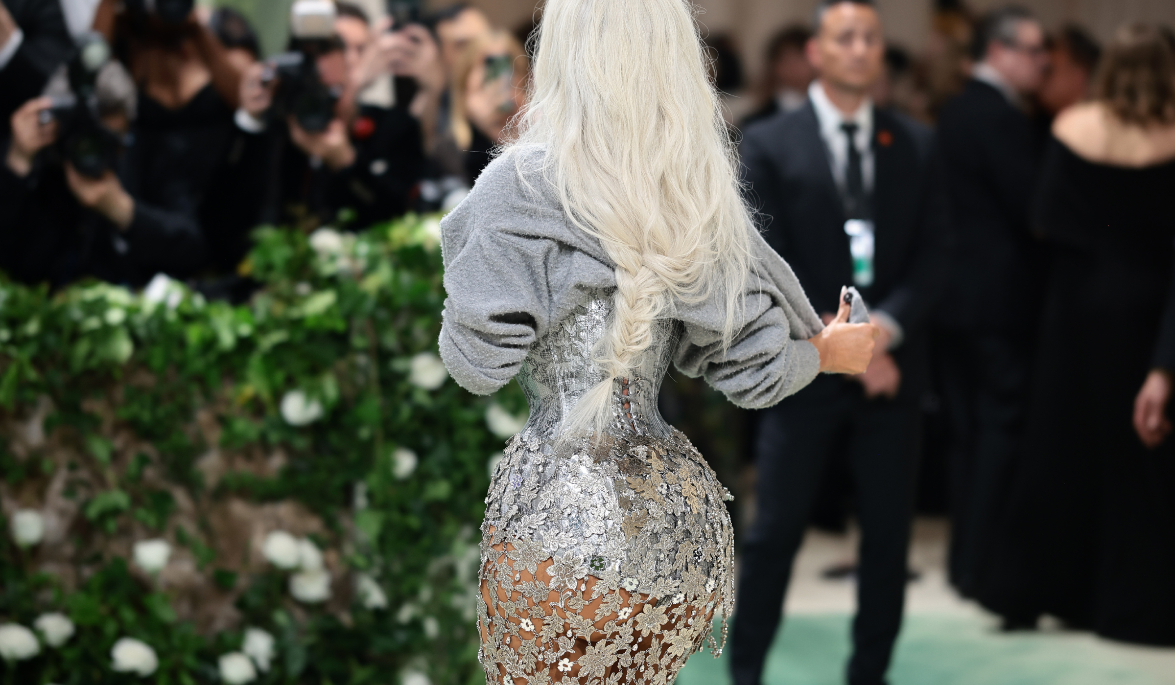Kim Kardashian, seen from behind, in a silver floral-embellished corset gown with a gray sweater over her shoulders and a long platinum braid.