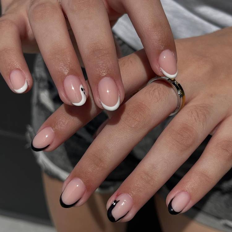 Short, rounded nails with a sheer pink base. One hand has thin white tips with a tiny ghost on one nail, and the other has thin black tips with a small black cat on one nail.