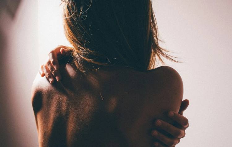 Close-up of a woman's bare shoulders and back from behind, softly backlit, with both hands crossed over her chest and shoulders