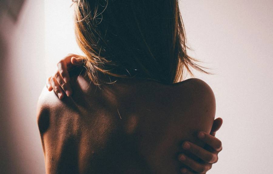 Close-up of a woman's bare shoulders and back from behind, softly backlit, with both hands crossed over her chest and shoulders