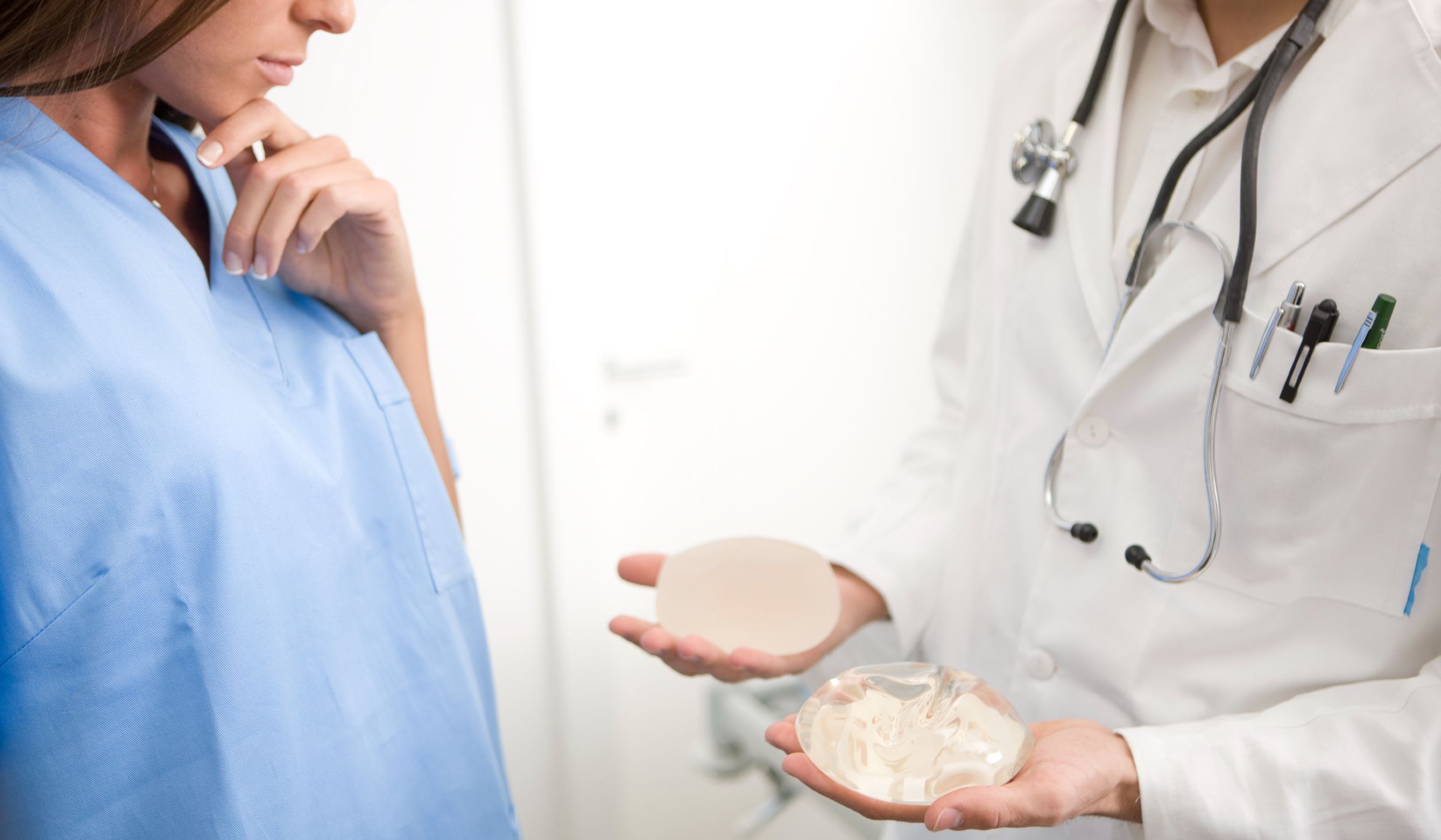 A doctor holds two different breast implant sizes in their palms while a patient looks on, considering her options during a consultation.