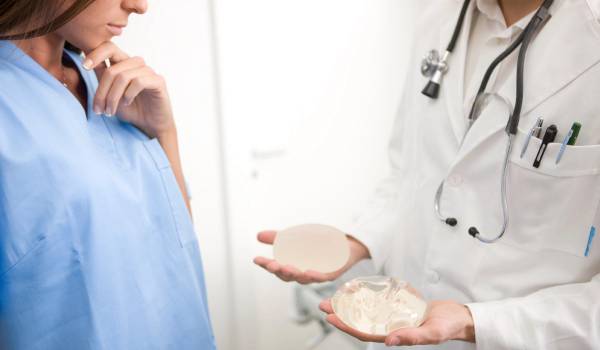 A doctor holds two different breast implant sizes in their palms while a patient looks on, considering her options during a consultation.