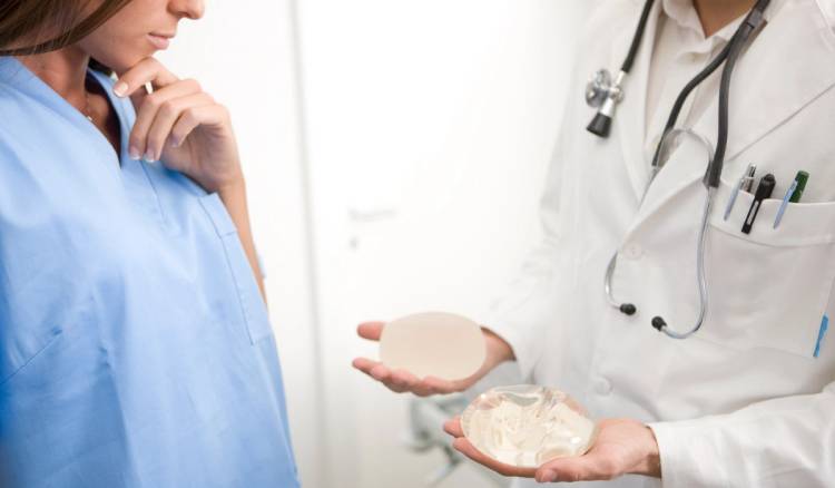 A doctor holds two different breast implant sizes in their palms while a patient looks on, considering her options during a consultation.
