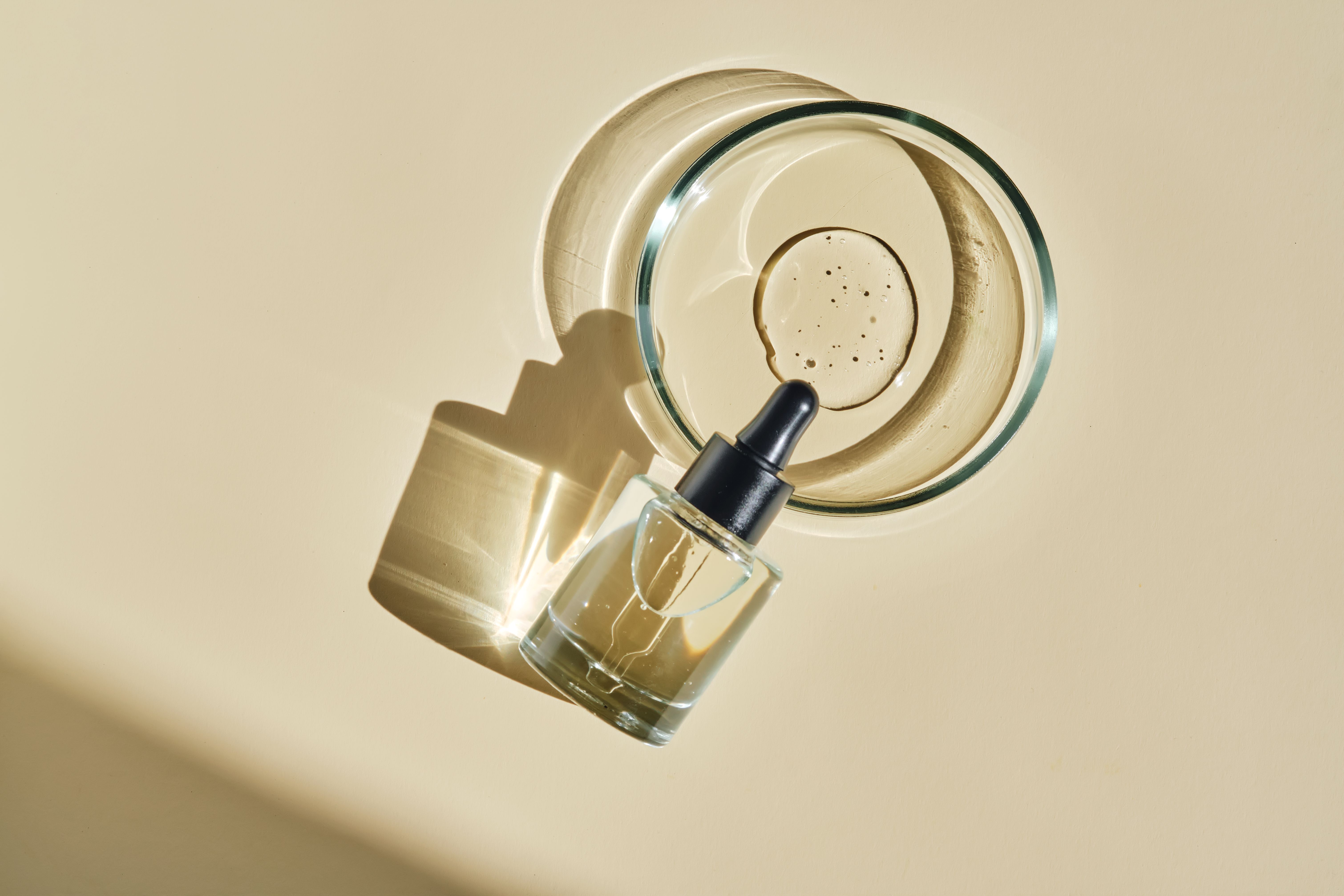 Overhead shot of a glass dropper bottle of facial serum resting against a shallow dish of skin-care product, both casting long shadows on a warm beige surface