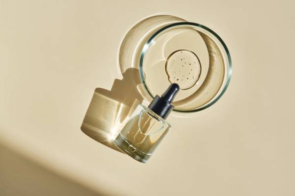 Overhead shot of a glass dropper bottle of facial serum resting against a shallow dish of skin-care product, both casting long shadows on a warm beige surface