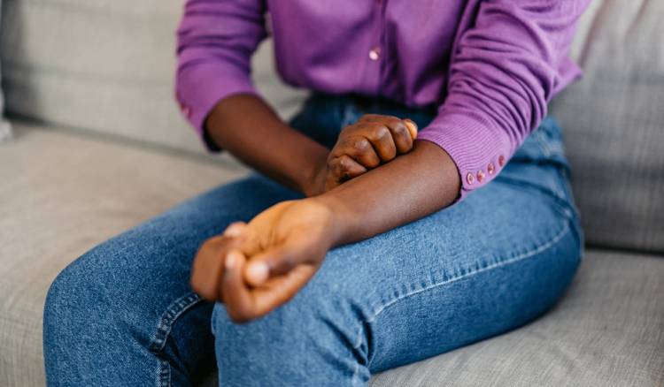 A person in a purple shirt scratches their forearm while sitting on a couch.
