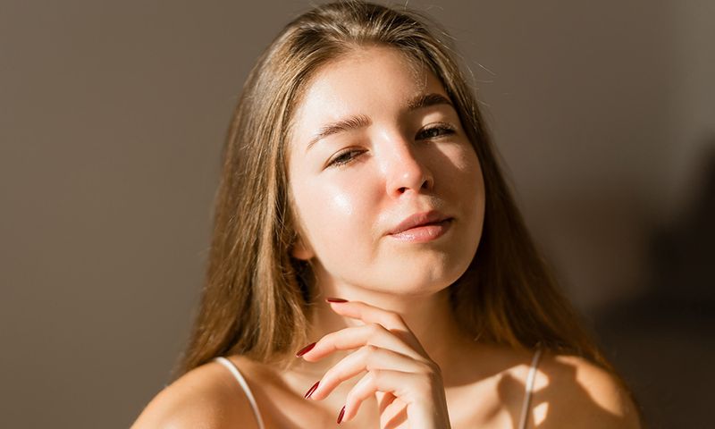 Portrait of a young woman touching her skin with her hand in the rays of the sun. Beautiful woman taking care of her skin, enjoying the home atmosphere. Beauty, skin care concept.
