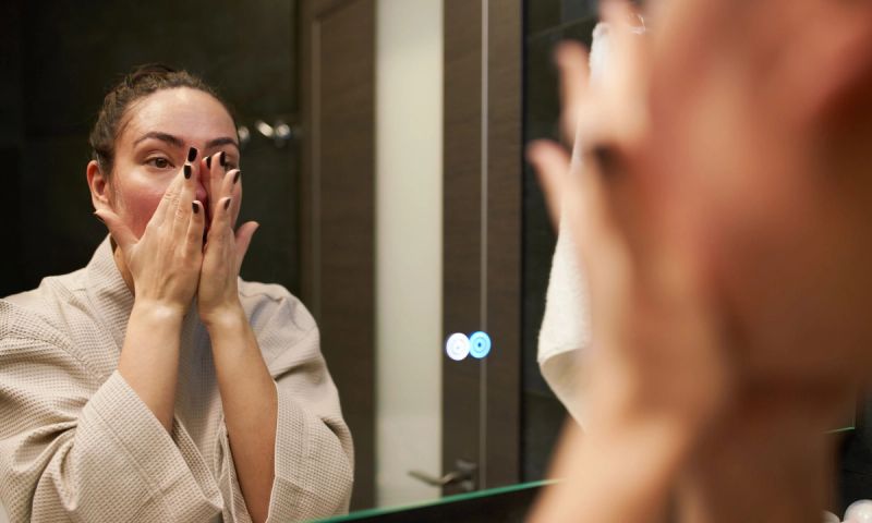 Female in beige bathrobe looking at mirror and massaging face during skin care routine in modern restroom.