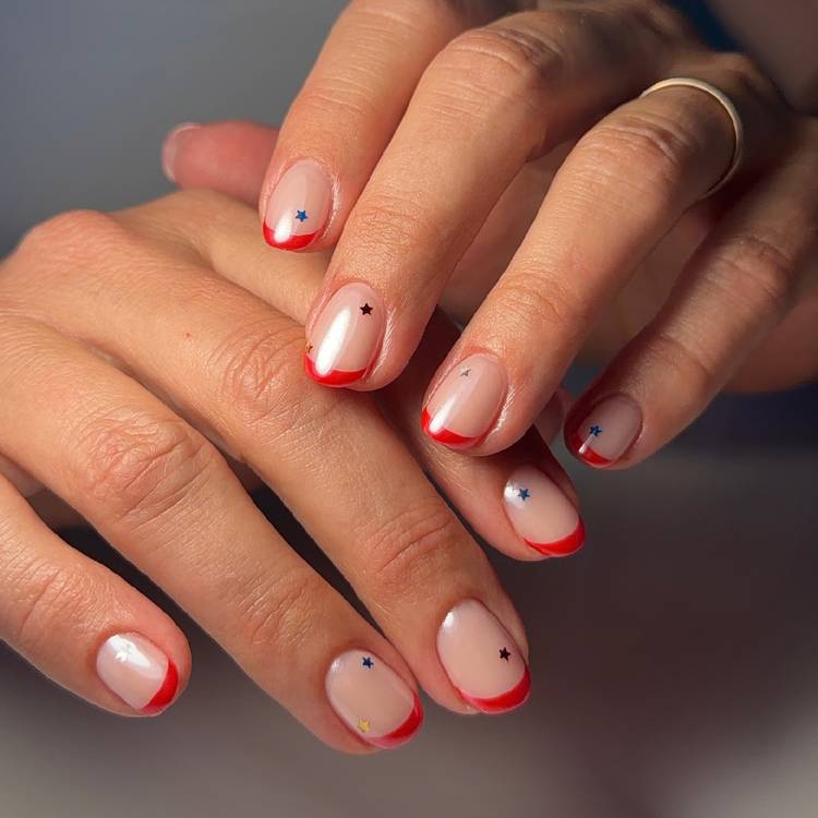 Close-up of hands with a red French tip manicure featuring small scattered star accents on a sheer nude base
