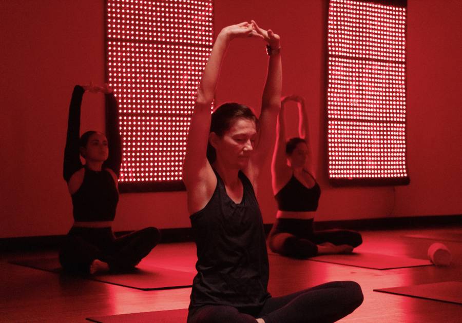 People in a yoga class stretch on mats in a dim studio illuminated by red light therapy panels mounted on the walls.