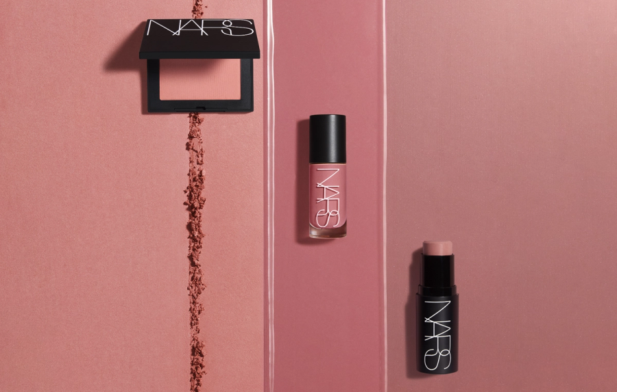 Three NARS blush formulas in a peach-pink shade—a powder compact, a liquid blush bottle and a cream stick—arranged on a rose background with a swipe of loose powder and a streak of liquid product