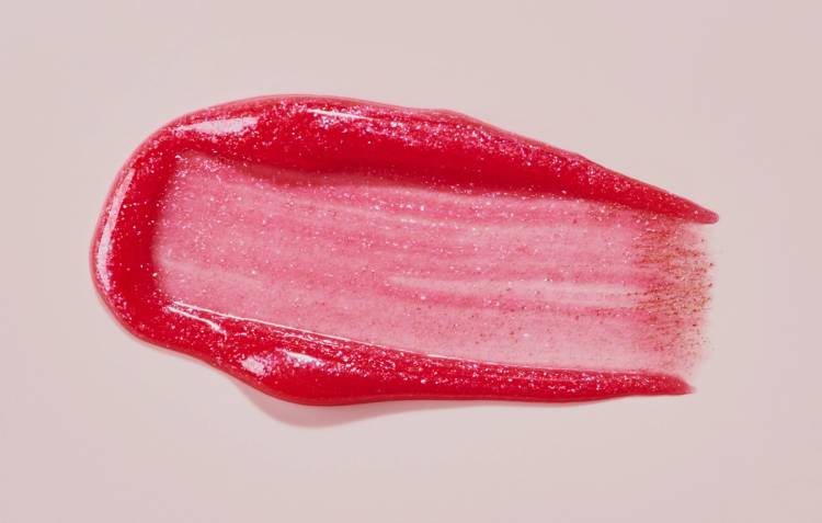 Close-up of a glittery red lip tint smeared in a swipe on a pink background, showing its glossy, sparkly texture