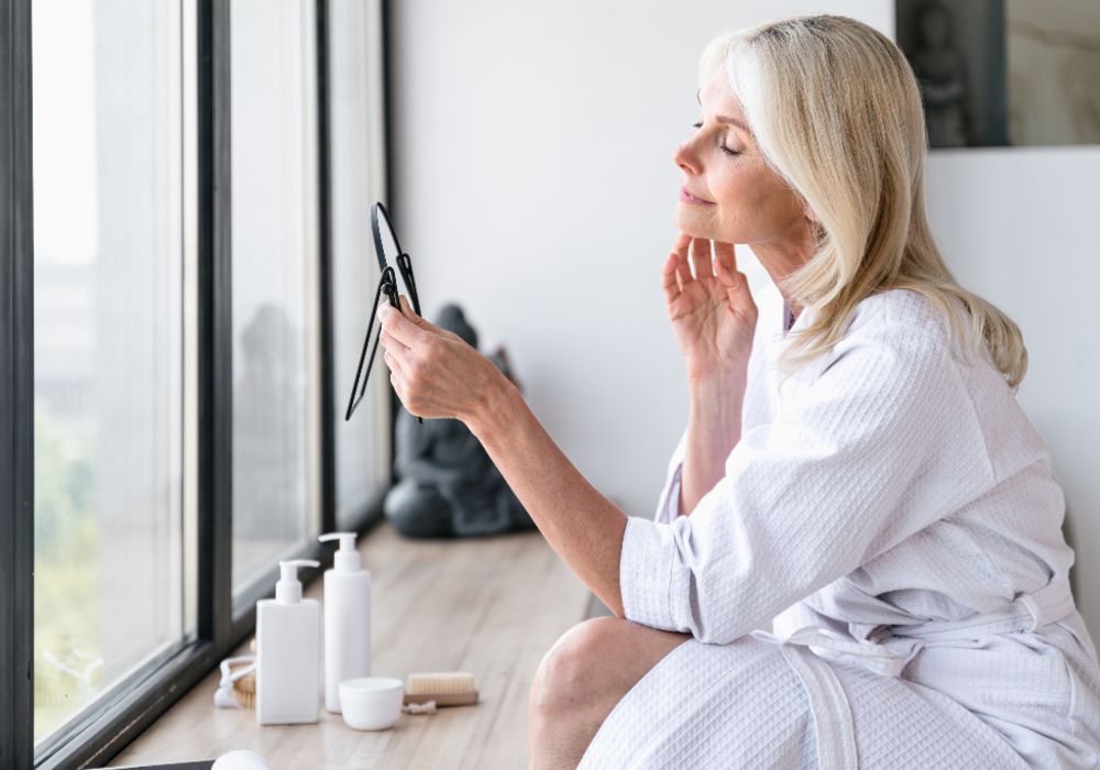 Woman examining her skin in a mirror by a window as part of a post-facelift skin care routine