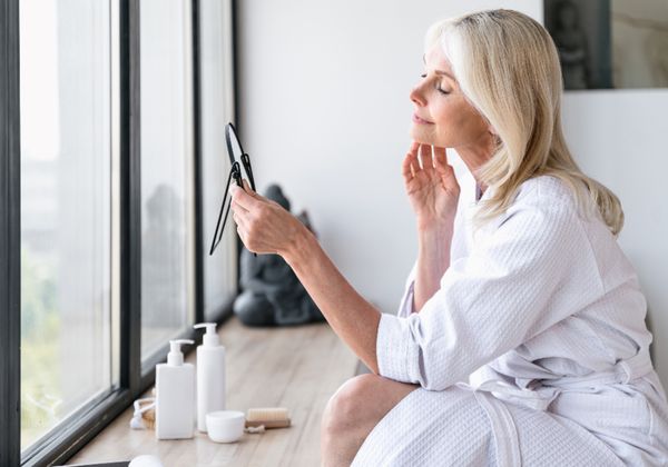 Woman examining her skin in a mirror by a window as part of a post-facelift skin care routine