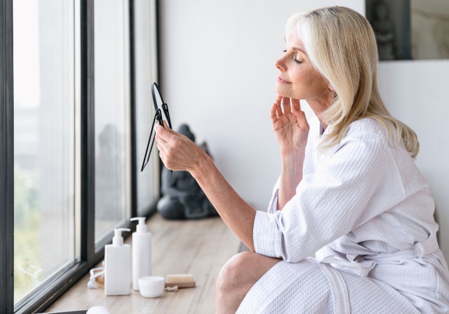 Woman examining her skin in a mirror by a window as part of a post-facelift skin care routine