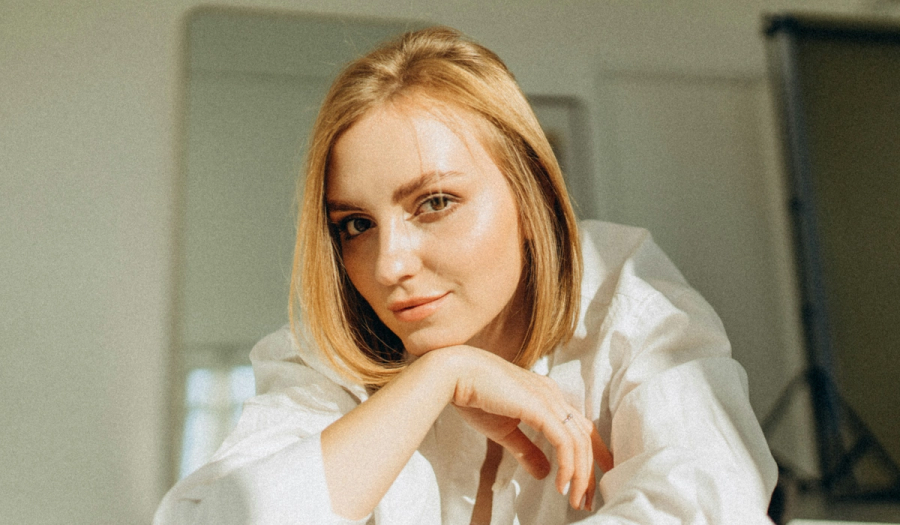 Woman with a blonde bob leans on her hand, resting her chin, with smooth, glowing skin and a relaxed expression, wearing a white button-down shirt.