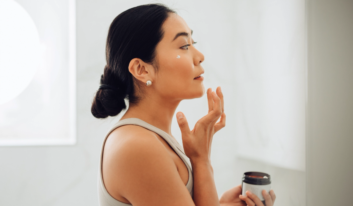 Woman applying face cream in a bright bathroom, shown in profile.
