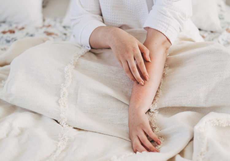 A person in a white long-sleeve shirt sits on a bed with cream-colored blankets, gently scratching irritated skin on their forearm.